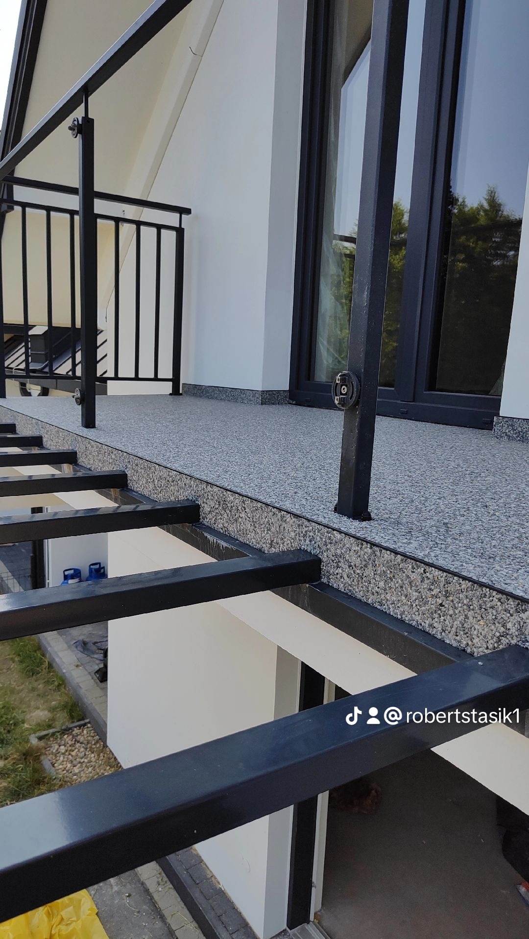 Szaro-biała, drobnoziarnista powierzchnia balkonu z czarną balustradą i ramą okienną, widok z góry na konstrukcję pergoli.