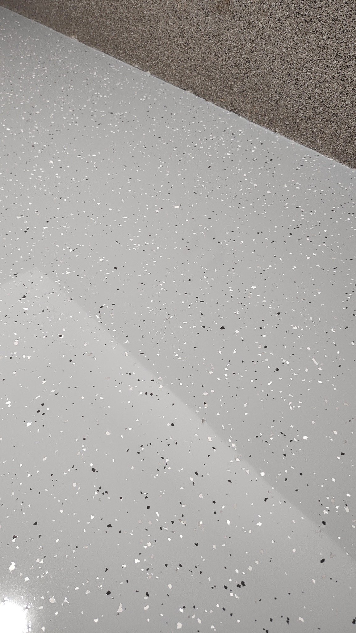 Jasnoszara powierzchnia podłogi z zatopionymi, błyszczącymi drobinkami w odcieniach bieli i szarości, widoczna krawędź ściany o chropowatej fakturze.