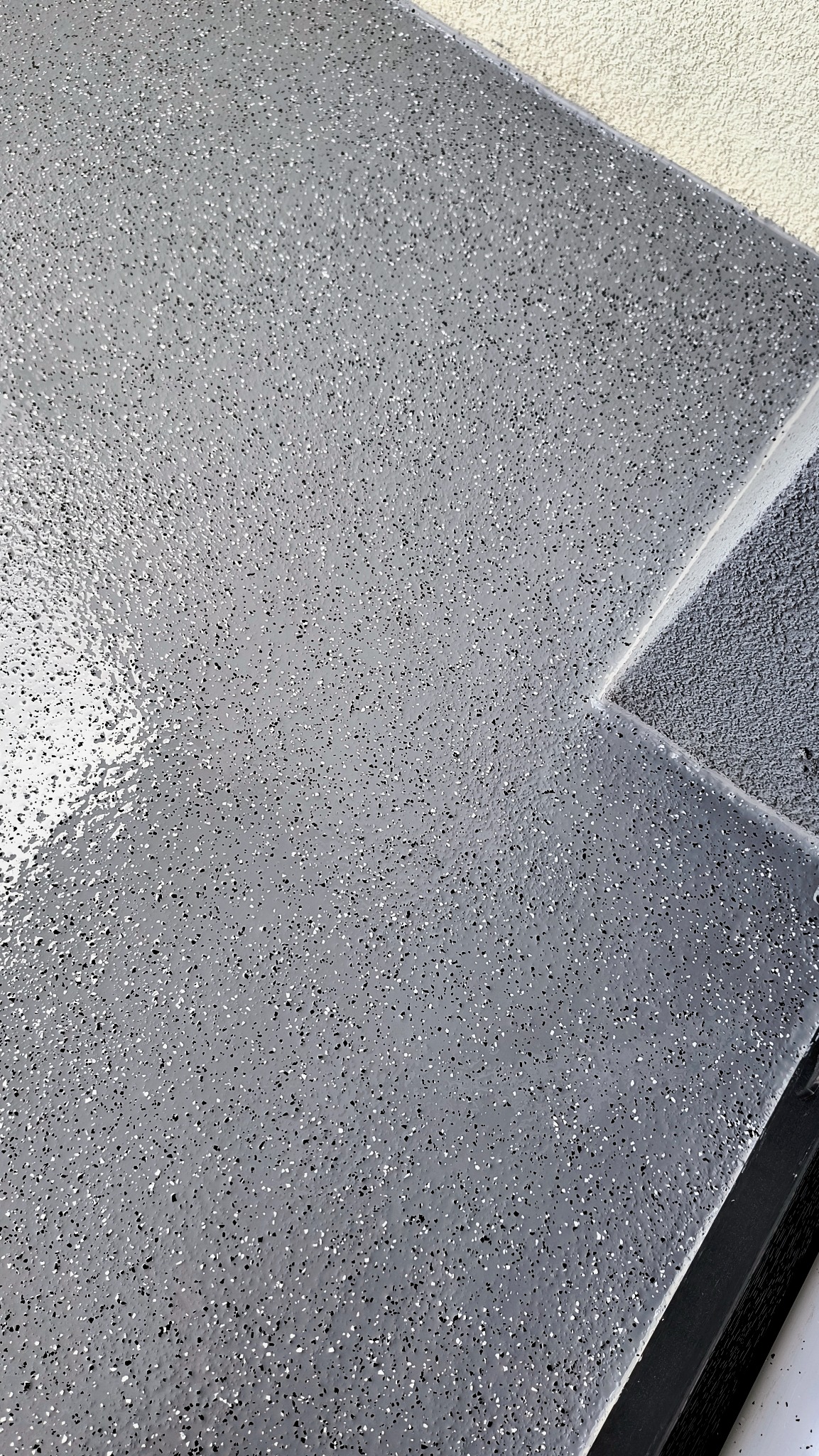 Jasnoszara, błyszcząca powierzchnia podłogi z drobnymi, czarnymi i białymi wtrąceniami, widoczna krawędź ściany i fragment ramy.