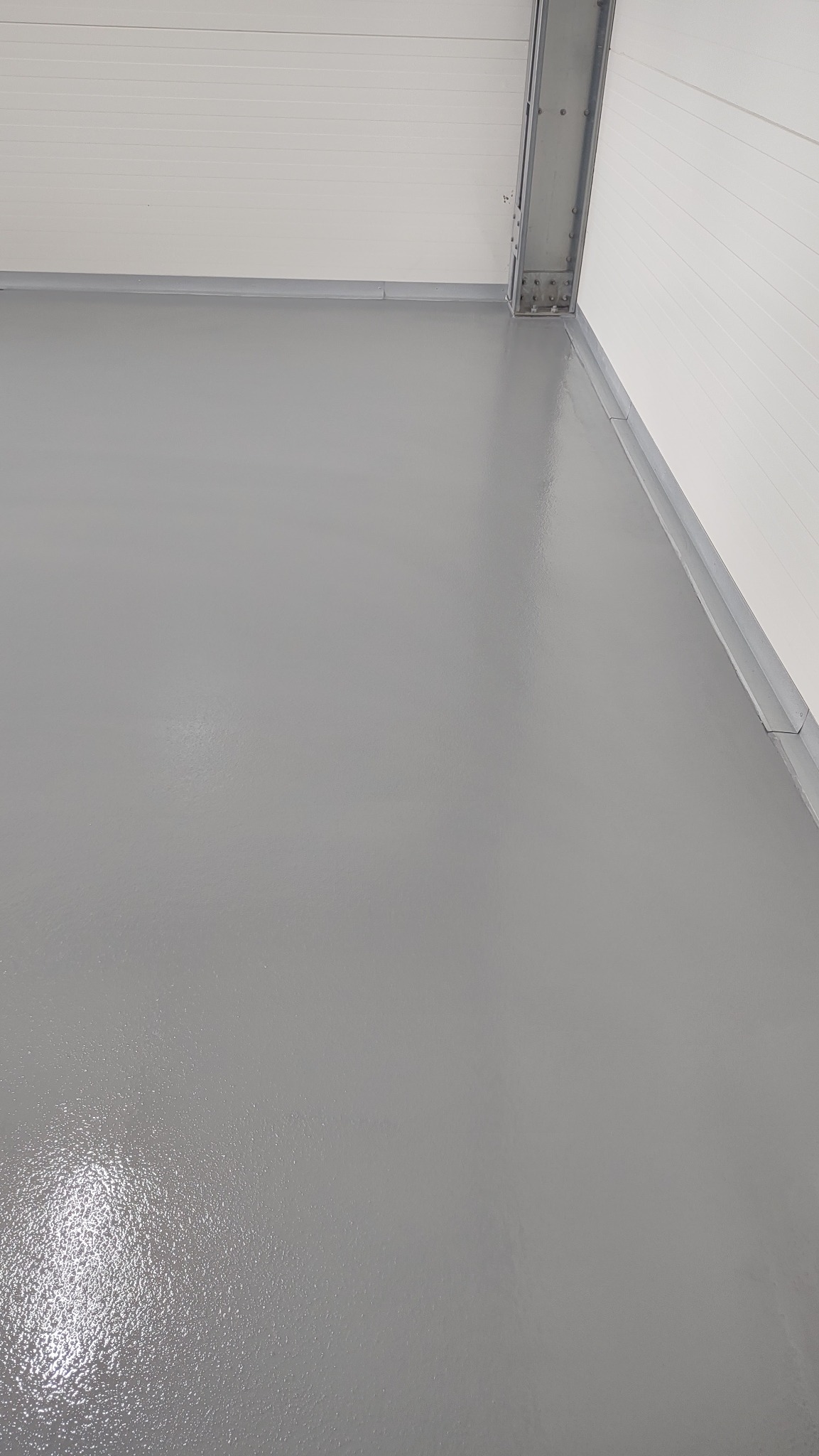 Świeżo wylana, gładka, szara podłoga w przemysłowym pomieszczeniu, widoczny metalowy profil na styku ściany i sufitu.