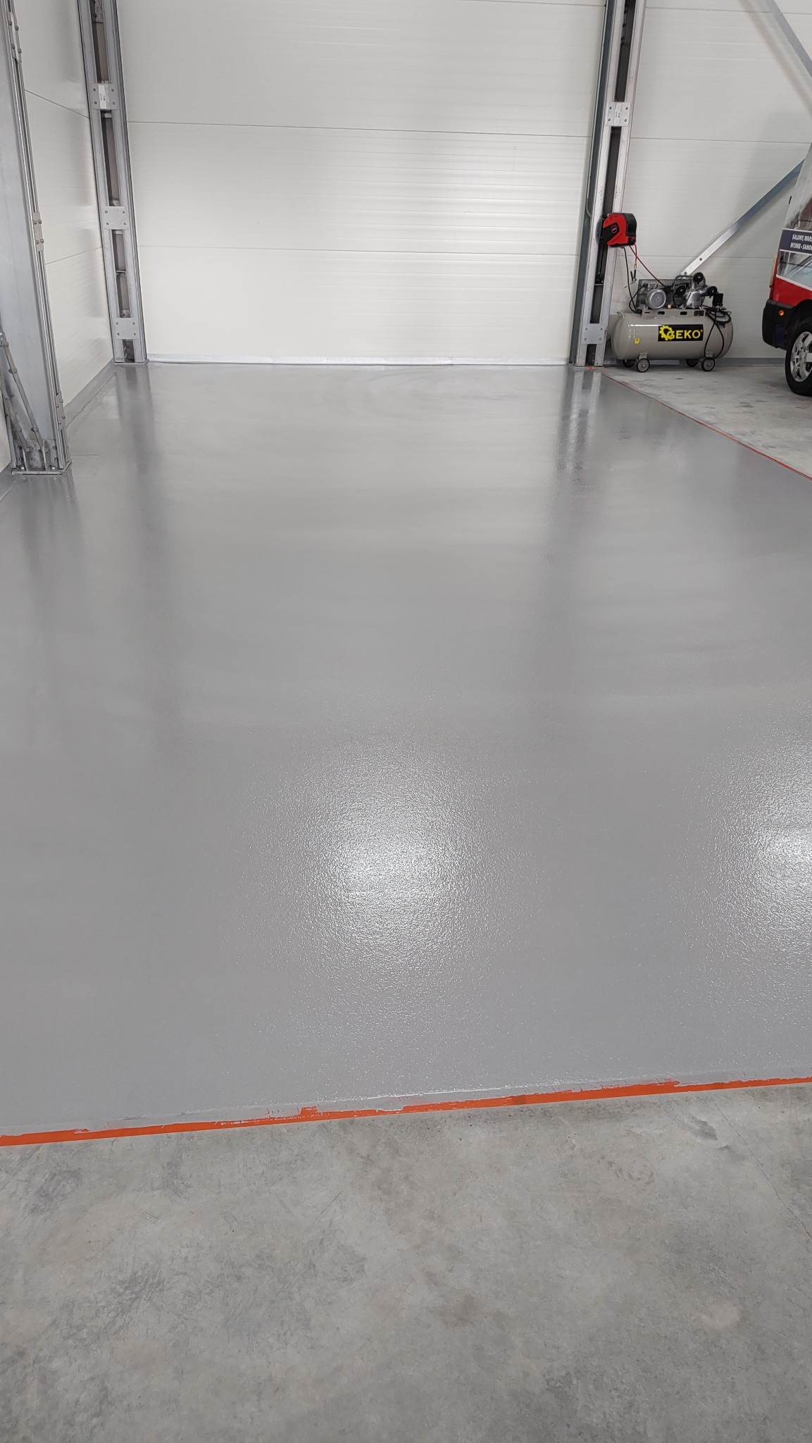 Gładka, szara powierzchnia w hali magazynowej, świeżo po wylaniu, z wyraźnie odznaczonymi pomarańczowymi liniami oddzielającymi od surowego betonu, kompresor powietrza w tle.