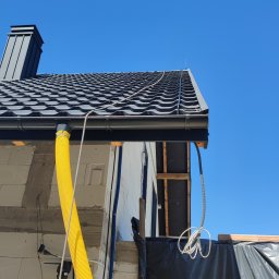 Grupa-Cyrek - Fragment dachu z czarną dachówką, rynną, kominem i instalacją odgromową. Widoczne elementy konstrukcji budynku w trakcie budowy.