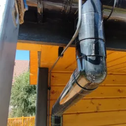 Zamontowany system rynnowy z czarną rurą spustową na drewnianej elewacji budynku, widok z dołu, fragment drabiny po lewej stronie kadru.