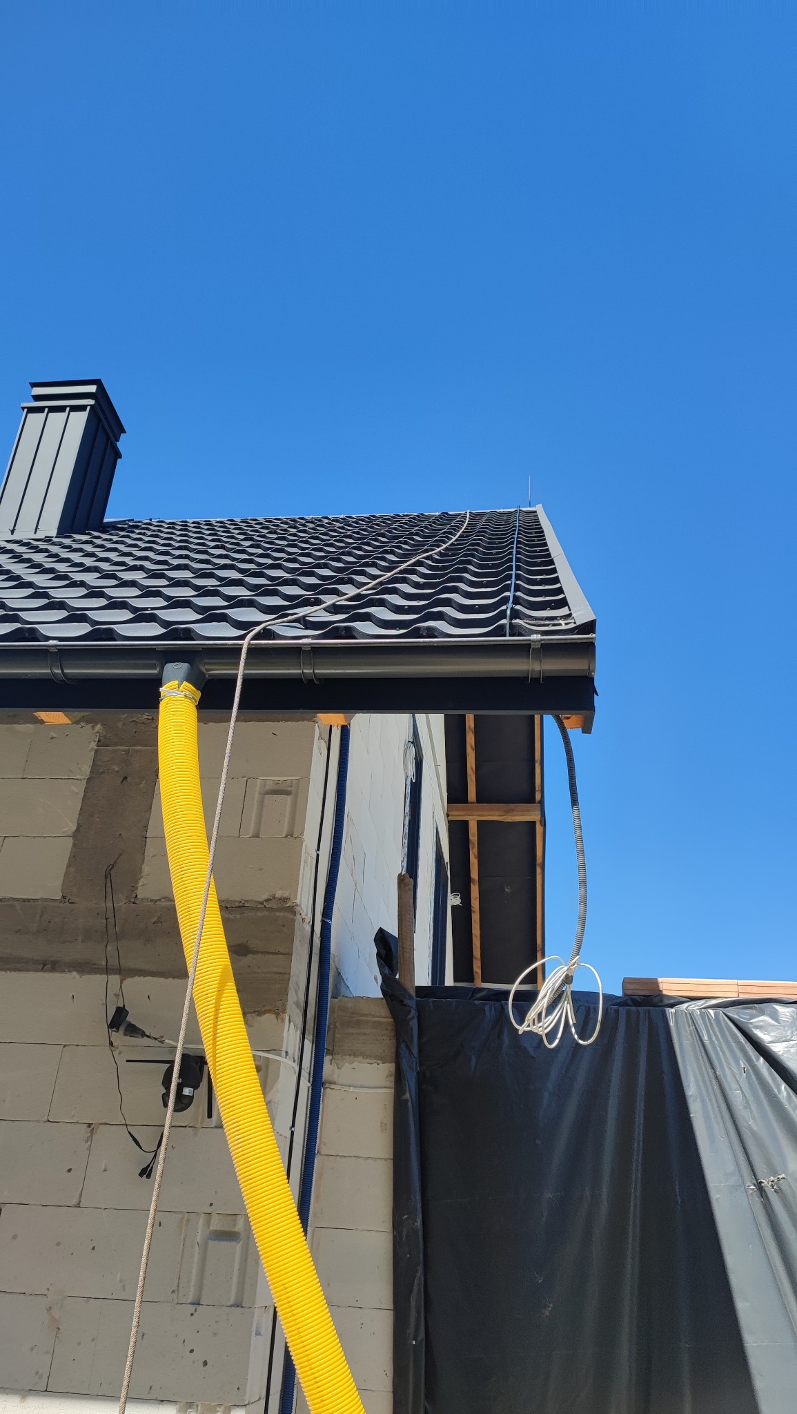 Fragment dachu z czarną dachówką, rynną, kominem i instalacją odgromową. Widoczne elementy konstrukcji budynku w trakcie budowy.