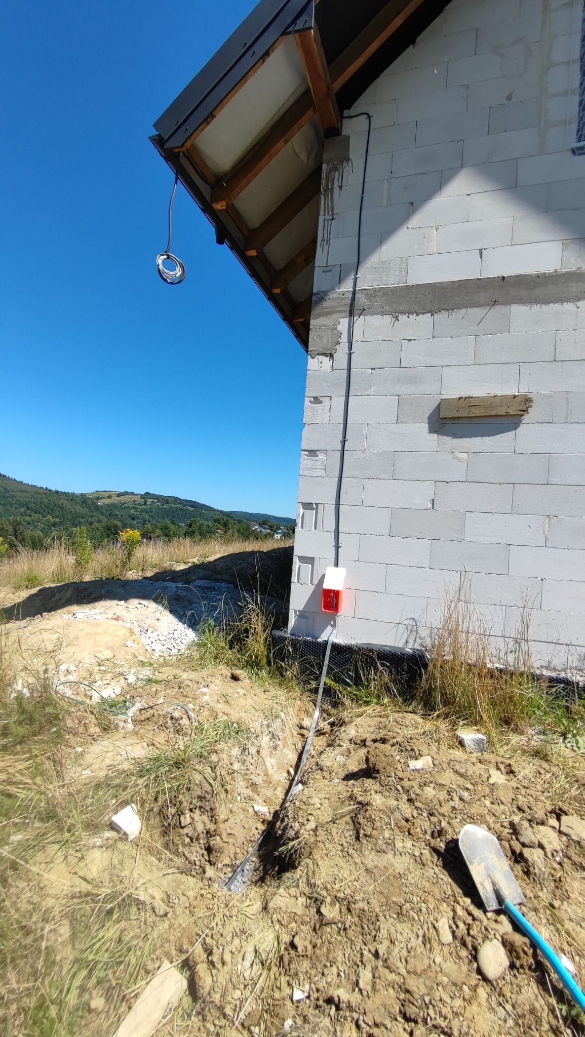 Biały budynek w trakcie budowy z zainstalowanym systemem odgromowym, widoczne przewody i skrzynka przyłączeniowa. Krajobraz zalesionych wzgórz w tle.