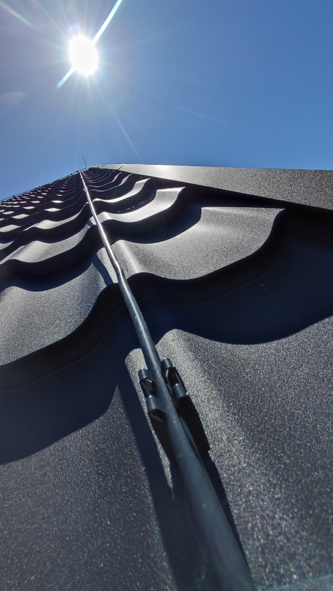 Metalowy pręt instalacji odgromowej na tle ciemnej blachodachówki w słoneczny dzień. Ujęcie od dołu podkreśla perspektywę i detale mocowania.