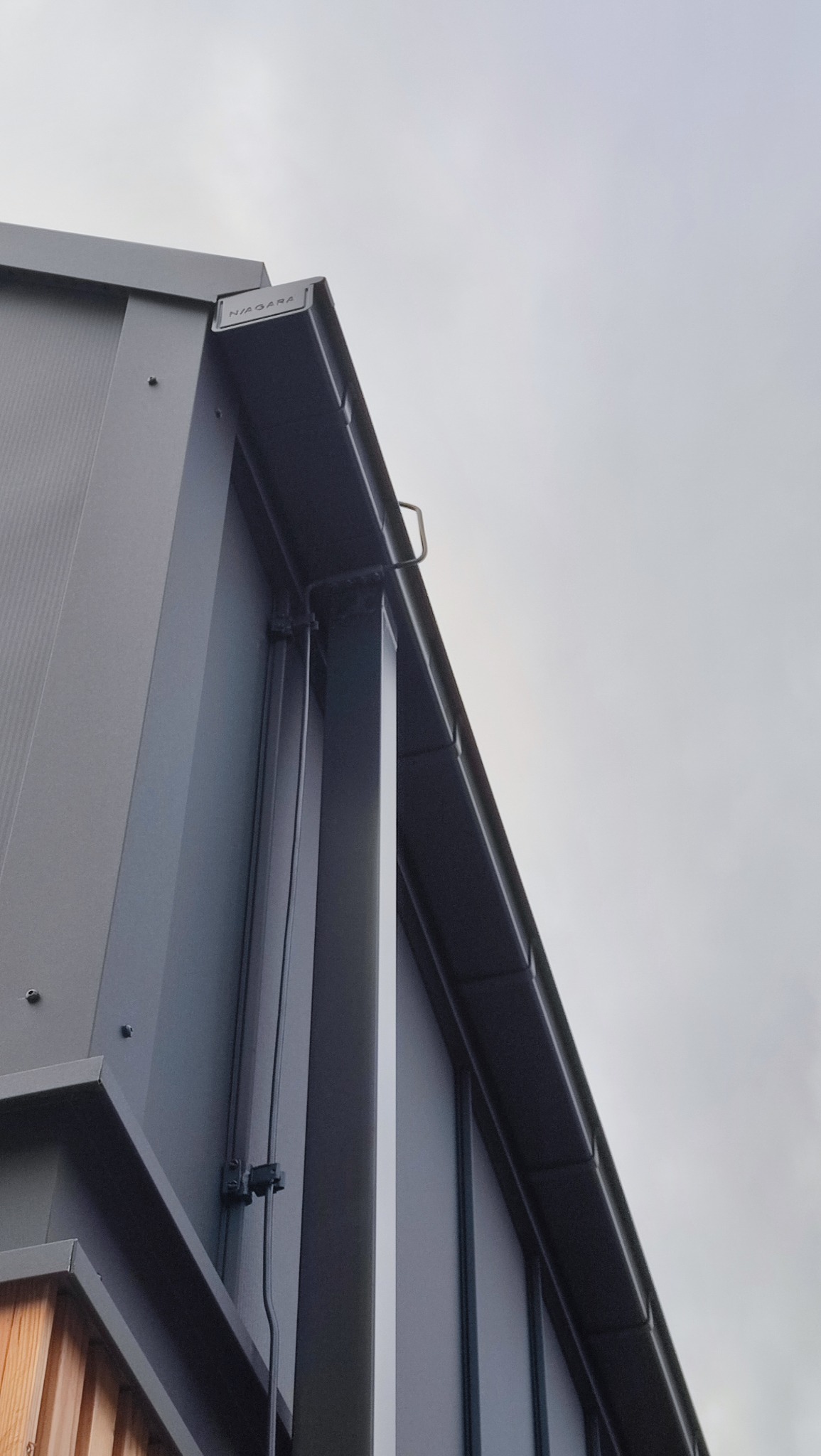 Szara, metalowa konstrukcja budynku z widocznym systemem rynnowym Niagara, uchwycona pod kątem z dołu na tle pochmurnego nieba.