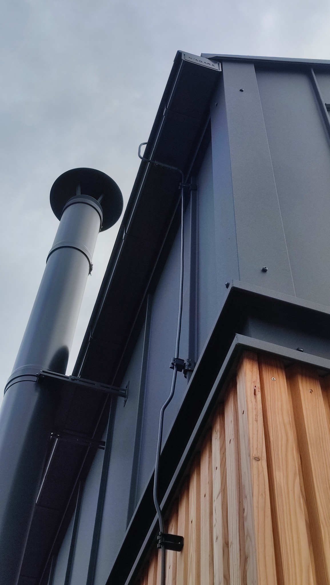Metalowy komin i pionowa elewacja budynku z drewnianą okładziną, widoczne przewody instalacji na tle pochmurnego nieba.