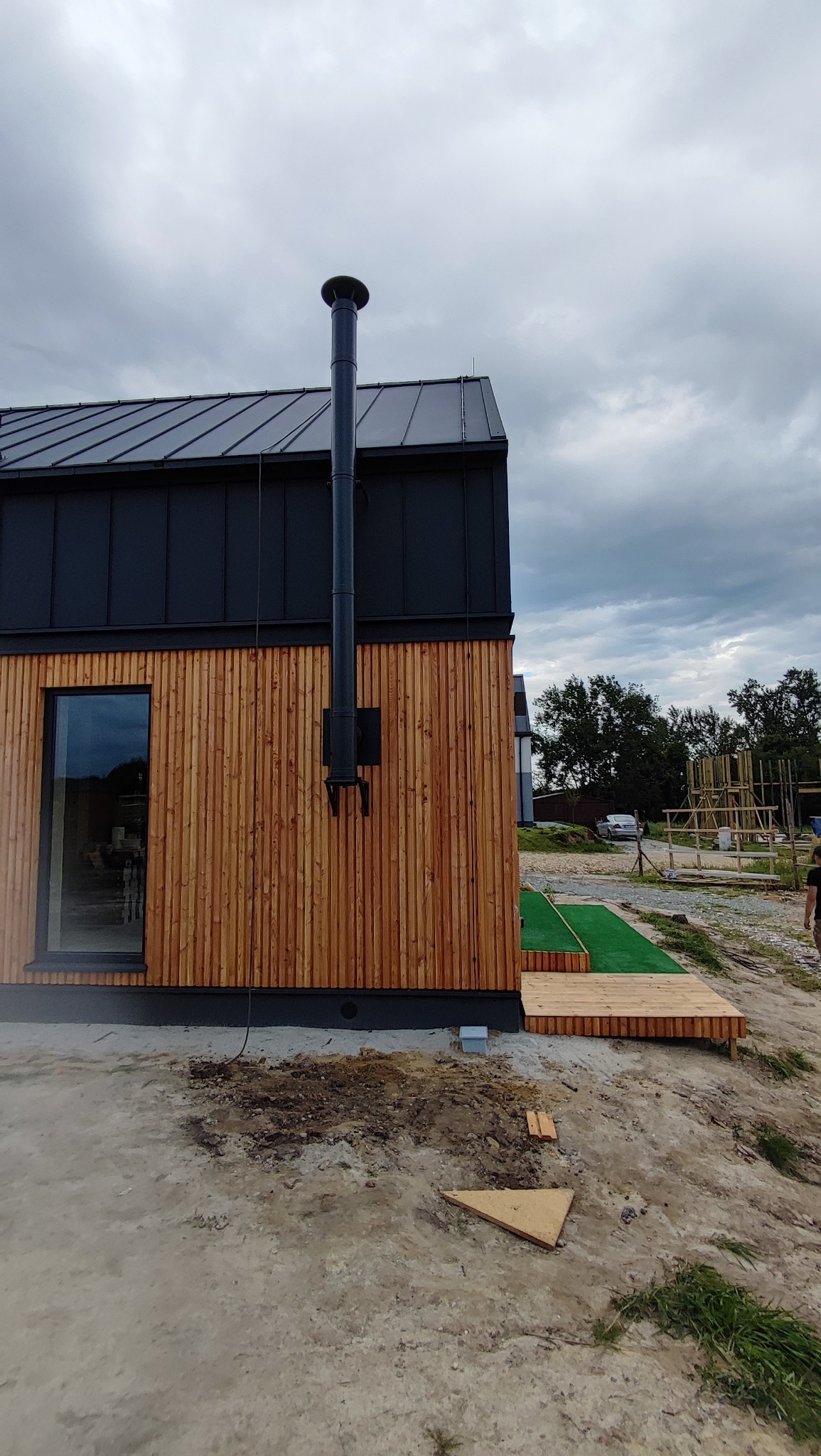 Nowoczesny dom z drewnianą elewacją i czarnym dachem z blachy. Widoczny komin, okno i fragment tarasu z zieloną wykładziną. Pochmurne niebo w tle.
