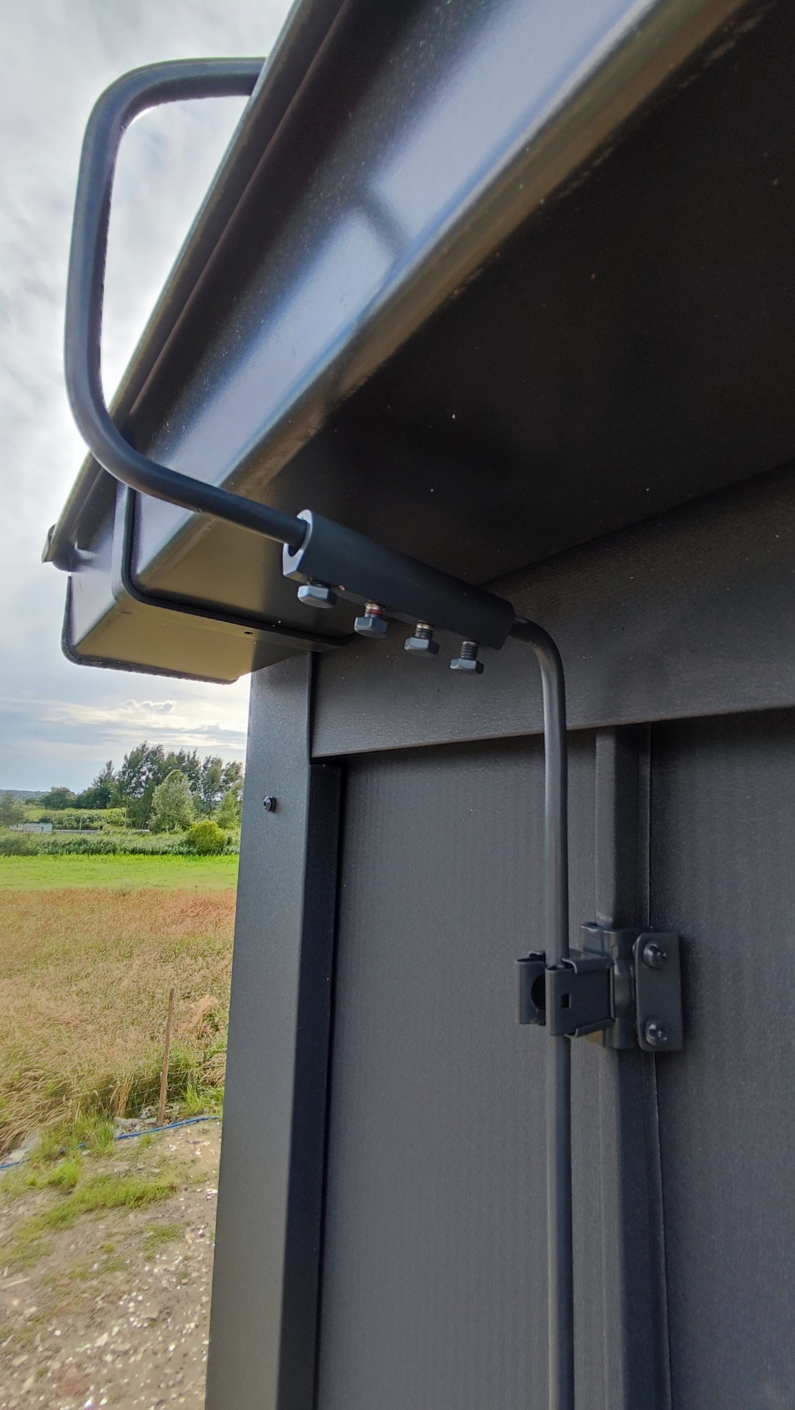 Detal montażu instalacji odgromowej na budynku z widokiem na łąkę w tle. Elementy mocujące i przewody w kolorze grafitowym.