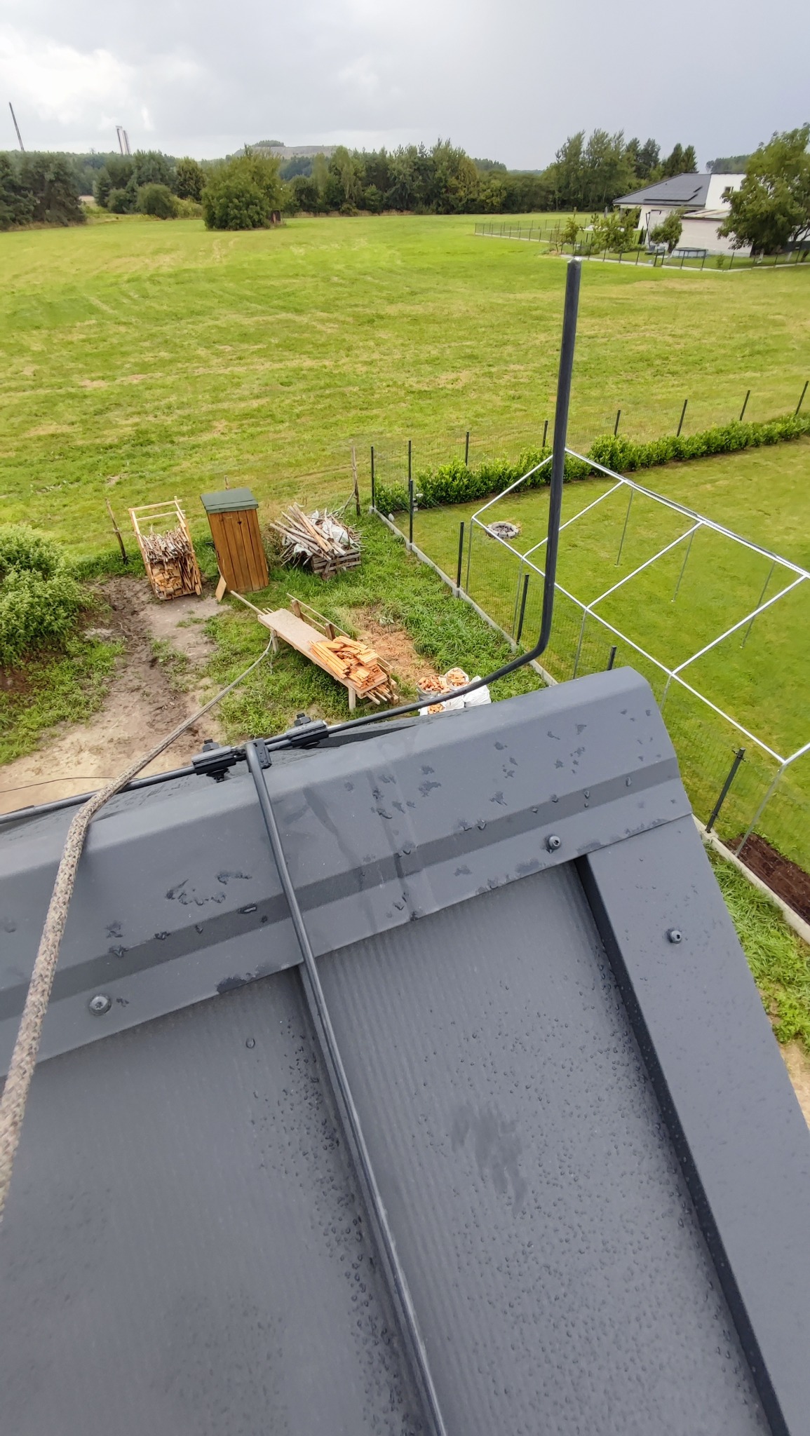 Szara blacha dachowa z zamontowaną instalacją odgromową. W tle widok na zielone pole, drzewa i zabudowania. Ujęcie z góry, po deszczu.