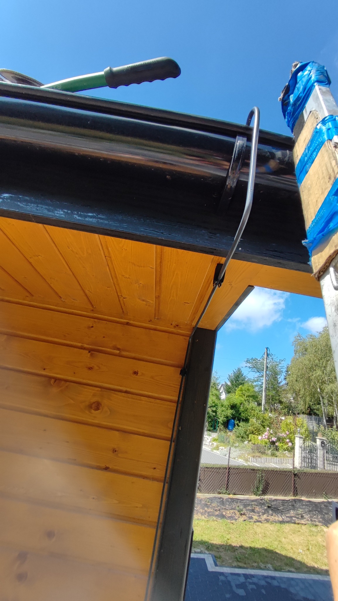 Montaż instalacji odgromowej na drewnianej konstrukcji dachu z użyciem drabiny. Widoczne narzędzia i elementy mocujące. Błękitne niebo w tle.