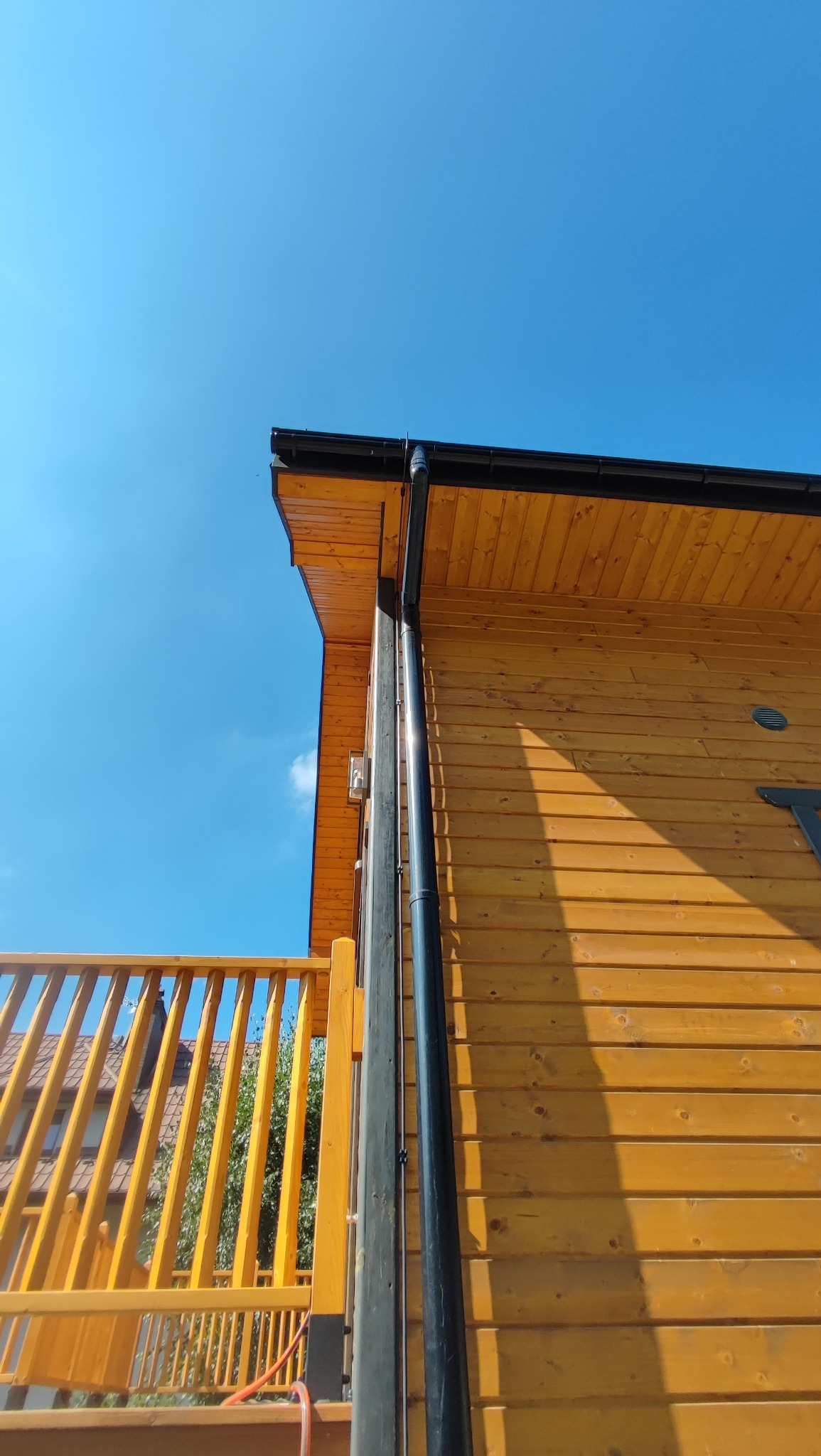 Drewniana elewacja domu z czarną rynną spustową i żółtą balustradą balkonu na tle błękitnego nieba w słoneczny dzień. Widok z dołu.