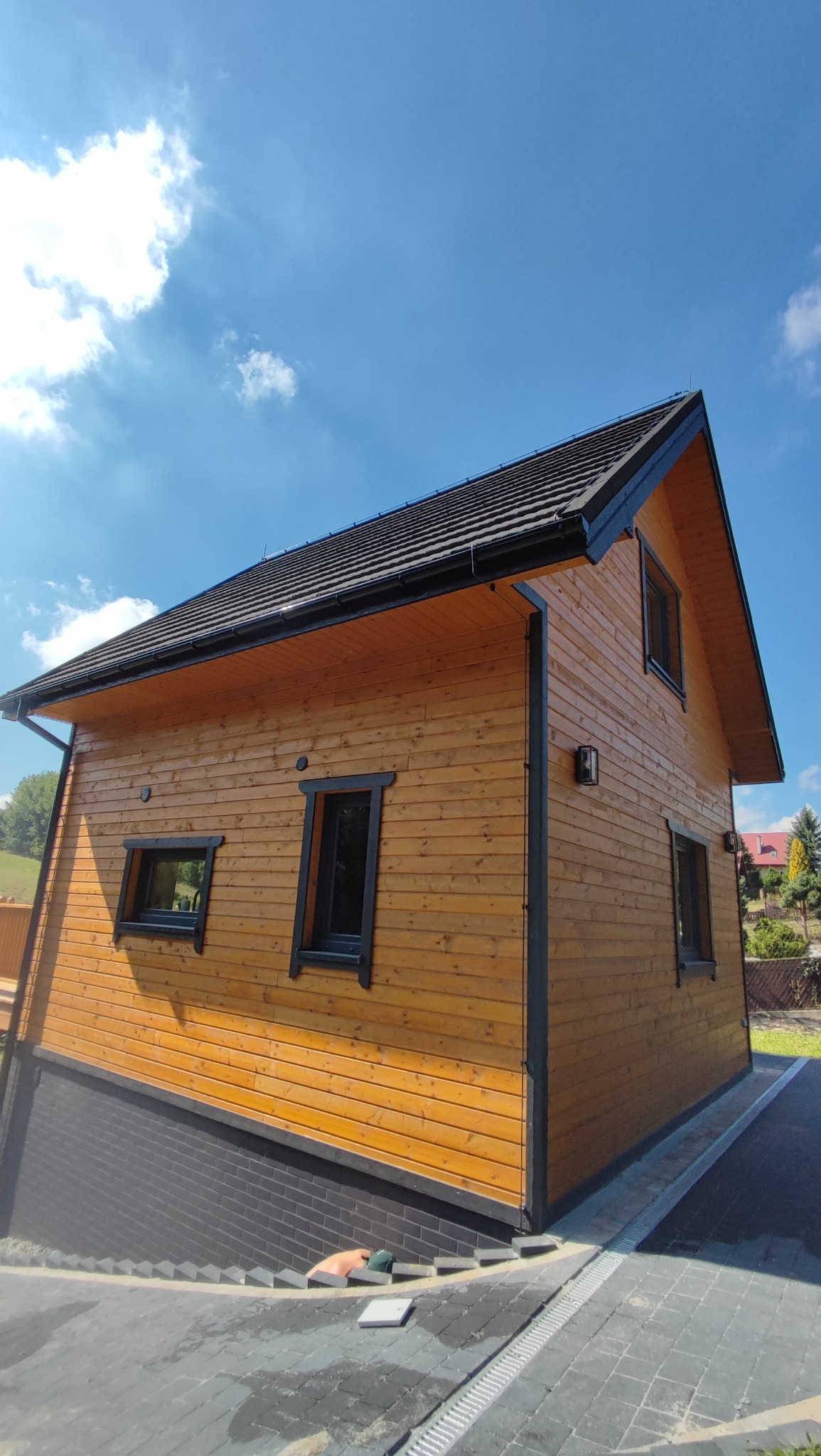 Drewniany dom z czarną stolarką okienną i dachem z dachówki, widziany pod kątem. Widoczne błękitne niebo i fragment zieleni w tle. Dom z elewacją z jasnego drewna.