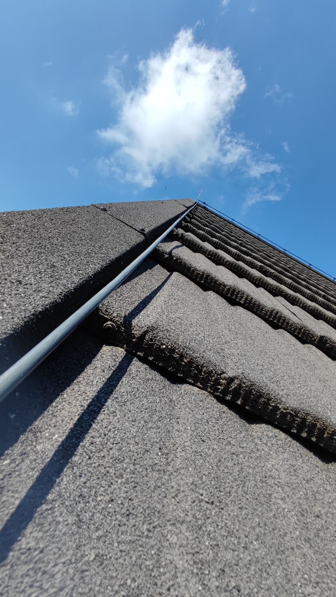 Szary dach pokryty dachówką bitumiczną z zamontowaną instalacją odgromową, na tle błękitnego nieba z pojedynczą chmurą. Ujęcie z dołu, podkreślające regularność ułożenia dachówek.
