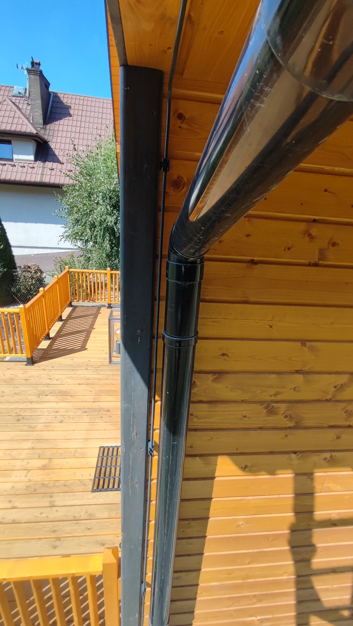 Zamontowana czarna rynna spustowa na drewnianej elewacji domu w Lubomierzu. Widoczny fragment tarasu z drewnianą balustradą i budynek w tle.