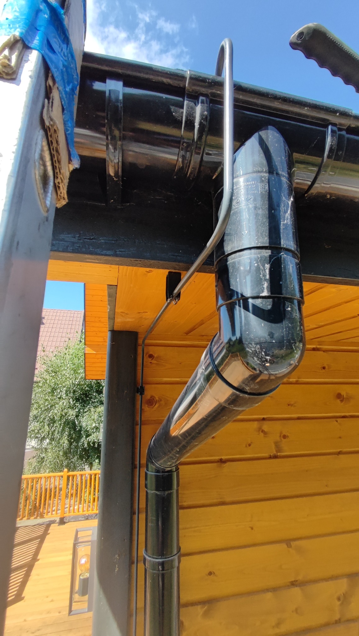 Zamontowany system rynnowy z czarną rurą spustową na drewnianej elewacji budynku, widok z dołu, fragment drabiny po lewej stronie kadru.