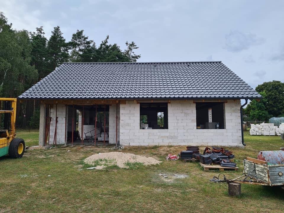 Dom w trakcie budowy z częściowo ukończonym dachem z szarej dachówki ceramicznej, widoczne pustaki, rusztowania, palety z dachówką i wózek widłowy w tle.