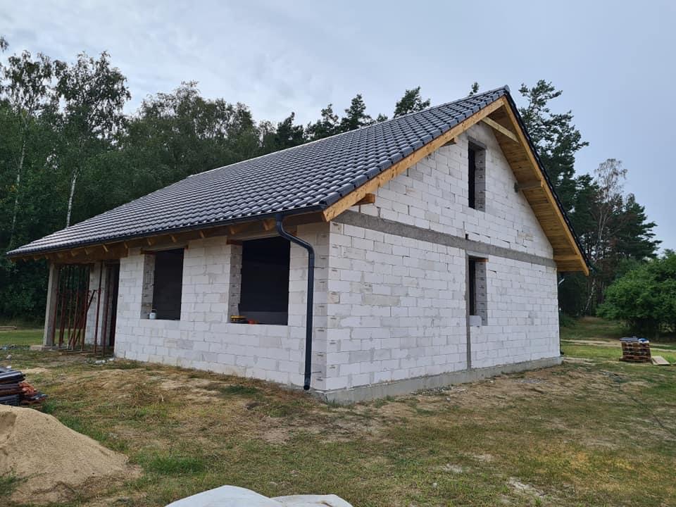 Surowy stan domu jednorodzinnego z szarej cegły, z zamontowaną czarną rynną i częściowo pokrytym dachem z ciemnej dachówki, widoczne elementy drewnianej więźby dachowej.