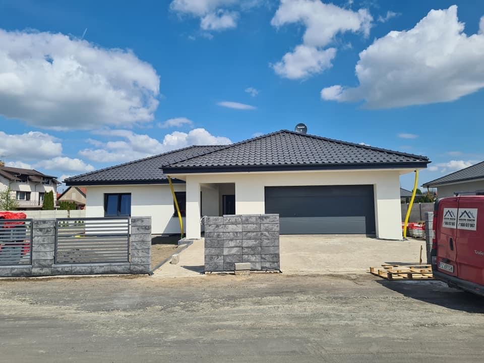 Nowoczesny dom jednorodzinny z ciemnoszarą dachówką ceramiczną, jasną elewacją i szarą bramą garażową, widoczny fragment ogrodzenia z betonowych bloczków, błękitne niebo z białymi obłokami w tle.