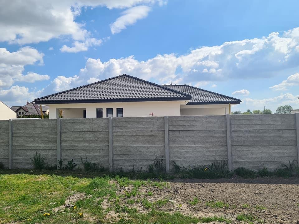 Nowoczesny dom jednorodzinny z ciemnoszarą dachówką, widoczny zza betonowego ogrodzenia panelowego w słoneczny dzień.