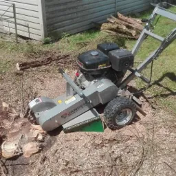 Frezarka do pni Jansen w trakcie pracy na posesji, widoczne świeże wióry po frezowaniu pnia, w tle sterta pociętych gałęzi i fragment budynku.