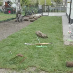 Układanie trawy z rolki na przygotowanym terenie, widoczne narzędzia ogrodnicze, rolki trawy i stos kostki brukowej w tle.