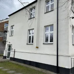 Jasna elewacja budynku z białymi oknami i czarną rynną, widoczny fragment chodnika z płyt imitujących pnie drzew i trawnik, w tle budynek z balkonami.