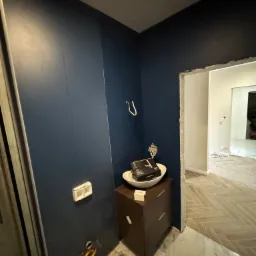 Niebieska ściana w łazience z przygotowanymi instalacjami, widoczne przyłącza wodno-kanalizacyjne, gładź na ścianach, płytki imitujące marmur na podłodze, szafka pod umywalkę z misą, w tle ułożona...