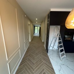 E-DOMIŚ - Długi korytarz z panelami ułożonymi w jodełkę, ściany w odcieniach beżu z dekoracyjnymi listwami, w tle kuchnia z marmurową podłogą i drabina oparta o ścianę.