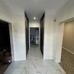 E-DOMIŚ - Korytarz w trakcie remontu z białymi ścianami, marmurową podłogą i widocznymi elementami instalacji elektrycznej. W głębi widoczny rower i szafa z przesuwnymi drzwiami.