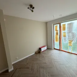 Pomalowane na beżowo wnętrze z parkietem ułożonym w jodełkę, okno zabezpieczone folią malarską, karton z lampą na podłodze.