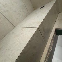 E-DOMIŚ - Ściana wyłożona beżowymi, matowymi płytkami ceramicznymi z ciemnymi fugami. Widoczny otwór w płytce. Krawędź ściany z widocznym klejem i fragmentem surowej ściany.