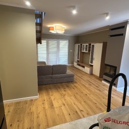 E-DOMIŚ - Jasne, przestronne wnętrze po remoncie: drewniana podłoga, szara sofa, białe meble. Widoczny fragment odkurzacza sugeruje trwające prace porządkowe.