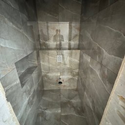 E-DOMIŚ - Wnętrze kabiny prysznicowej z szarymi płytkami imitującymi marmur, widok z dołu na sufit z odbiciem osoby robiącej zdjęcie. Widoczne niedokończone wykończenia.