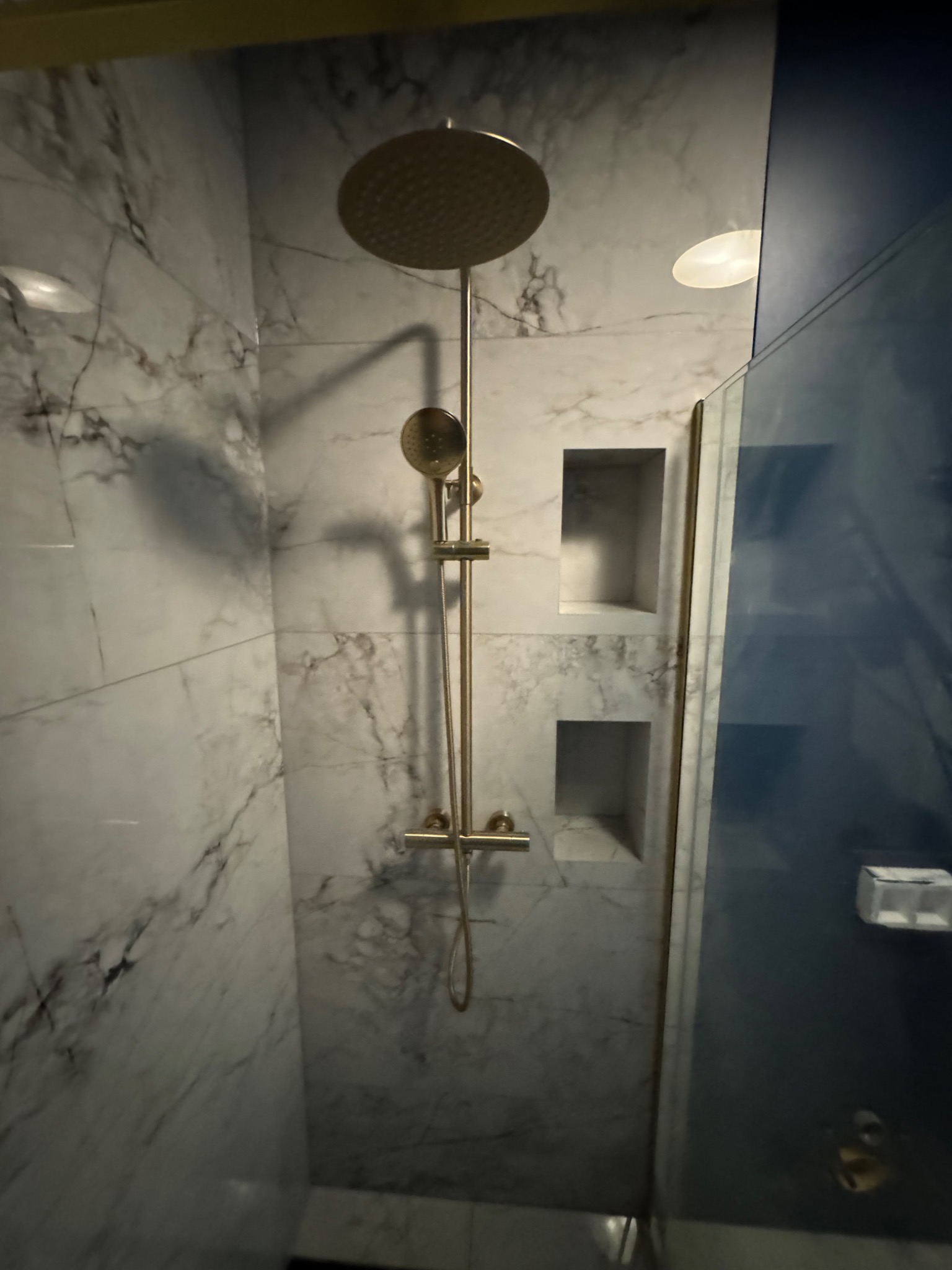 Nowoczesna kabina prysznicowa z marmurowymi płytkami, złotą armaturą i wnękami na kosmetyki.