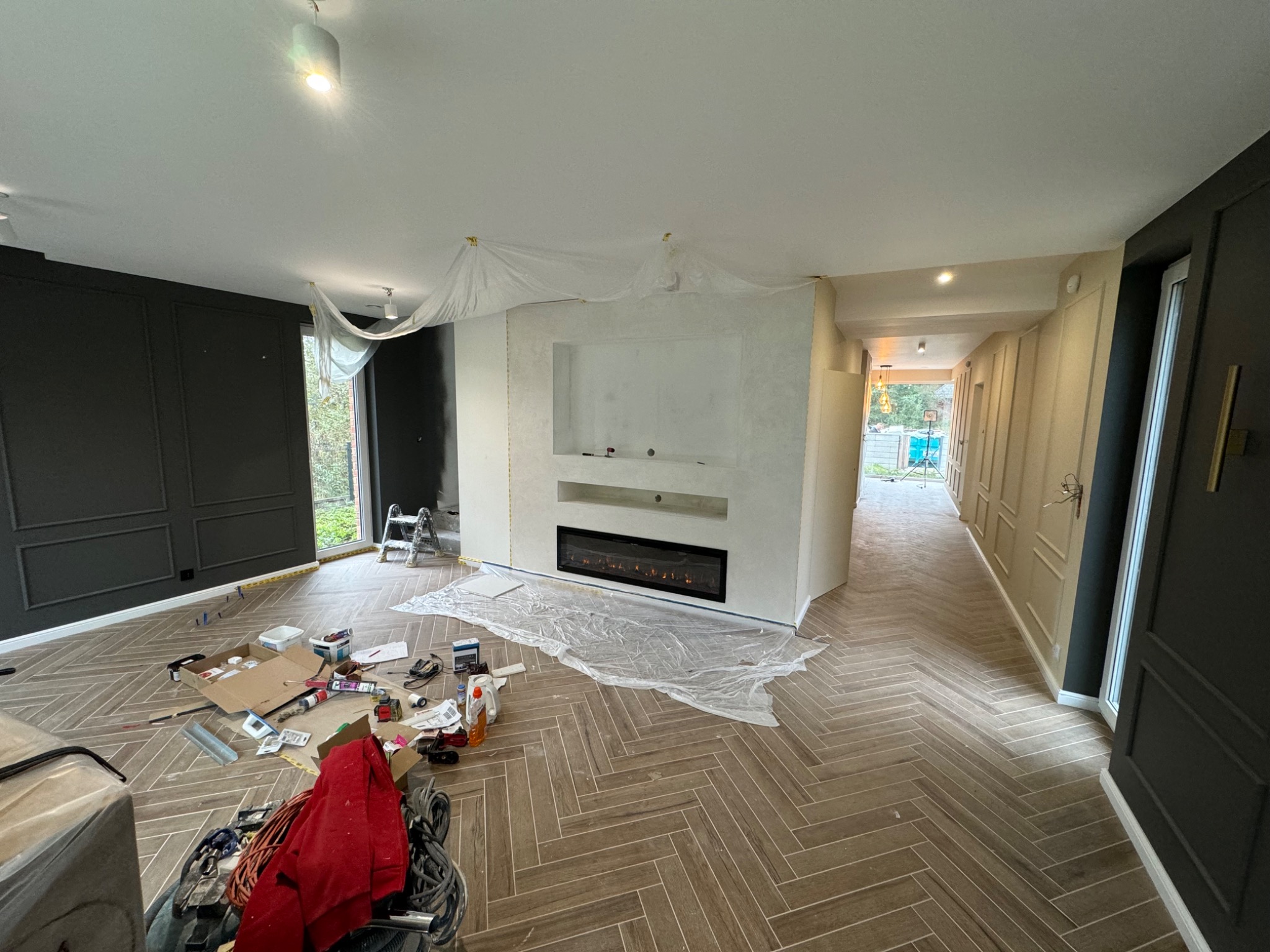 Salon w trakcie wykańczania: ułożona podłoga w jodełkę, ściany częściowo pomalowane, zabezpieczone folią, widoczne narzędzia i materiały budowlane.