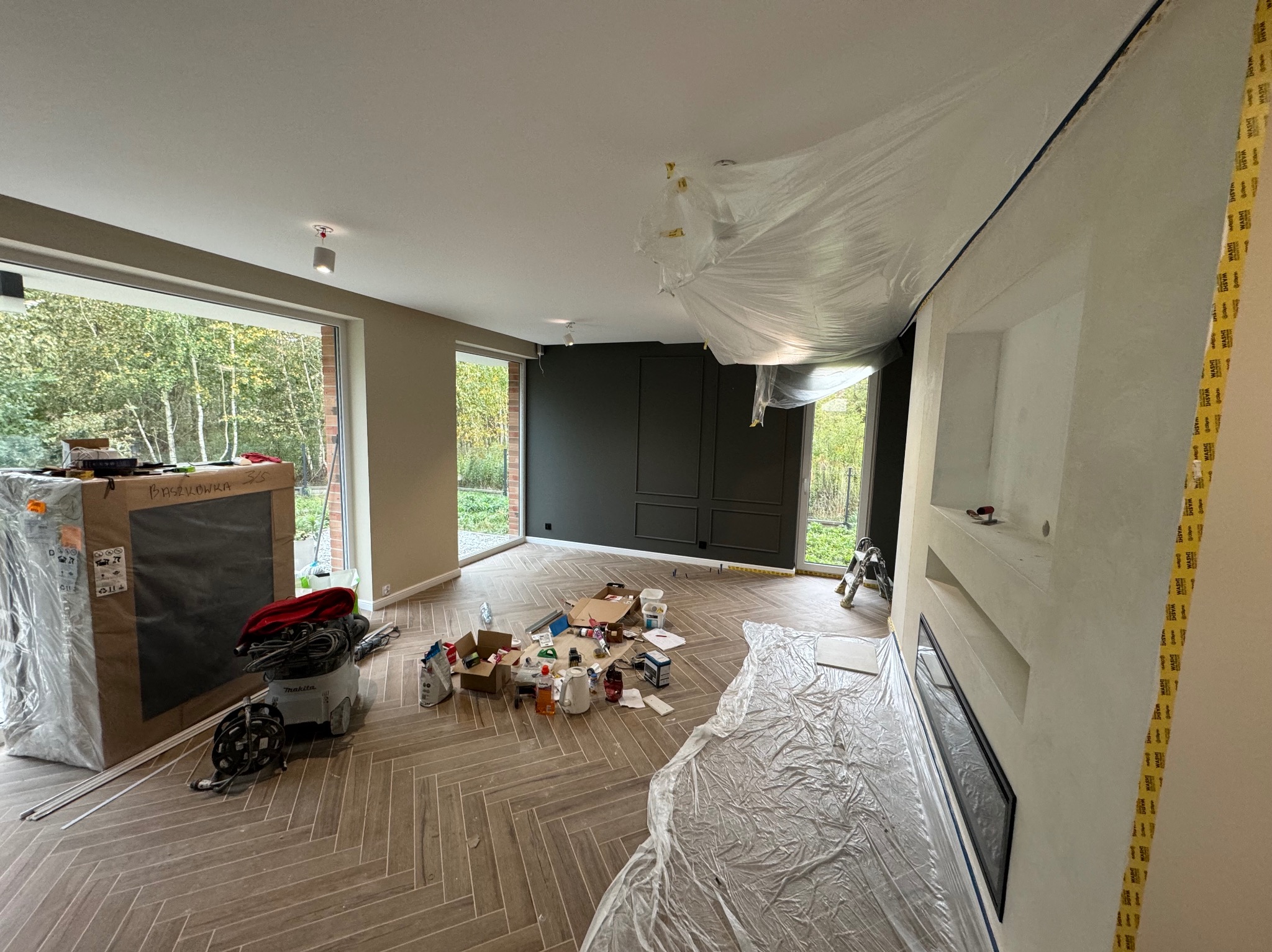 Pokój w trakcie remontu: podłoga z paneli ułożonych w jodełkę, ściany częściowo pomalowane na ciemny kolor, meble i podłoga zabezpieczone folią, widoczne narzędzia i puszki z farbą.