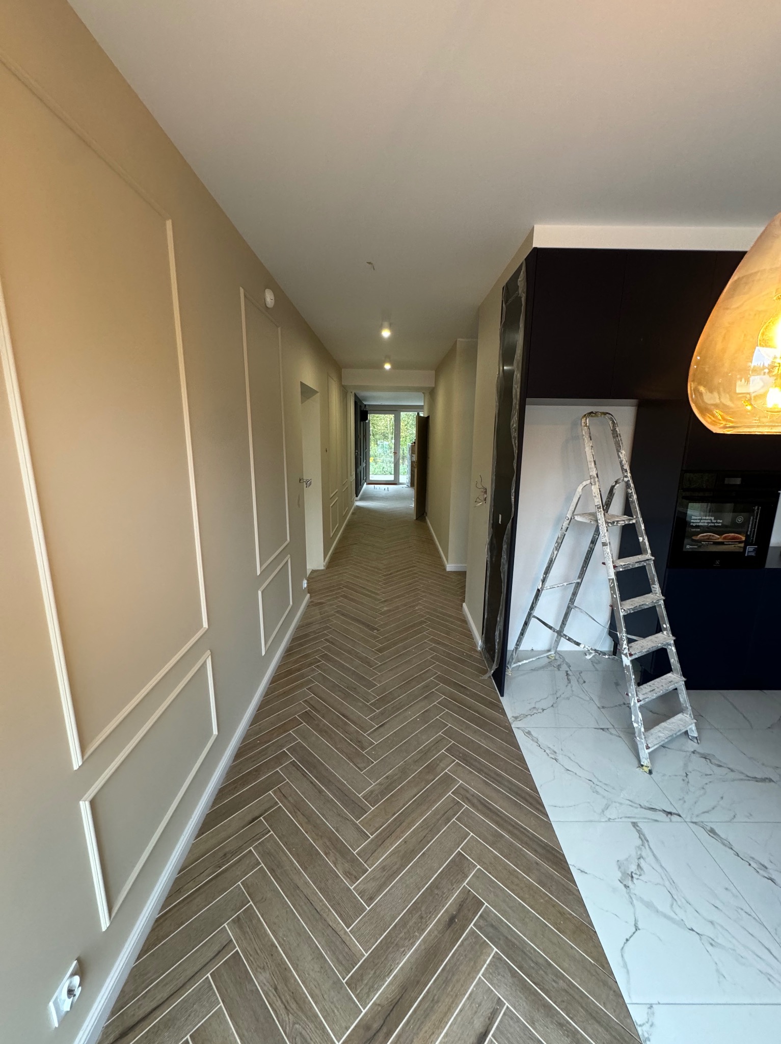 Długi korytarz z panelami ułożonymi w jodełkę, ściany w odcieniach beżu z dekoracyjnymi listwami, w tle kuchnia z marmurową podłogą i drabina oparta o ścianę.