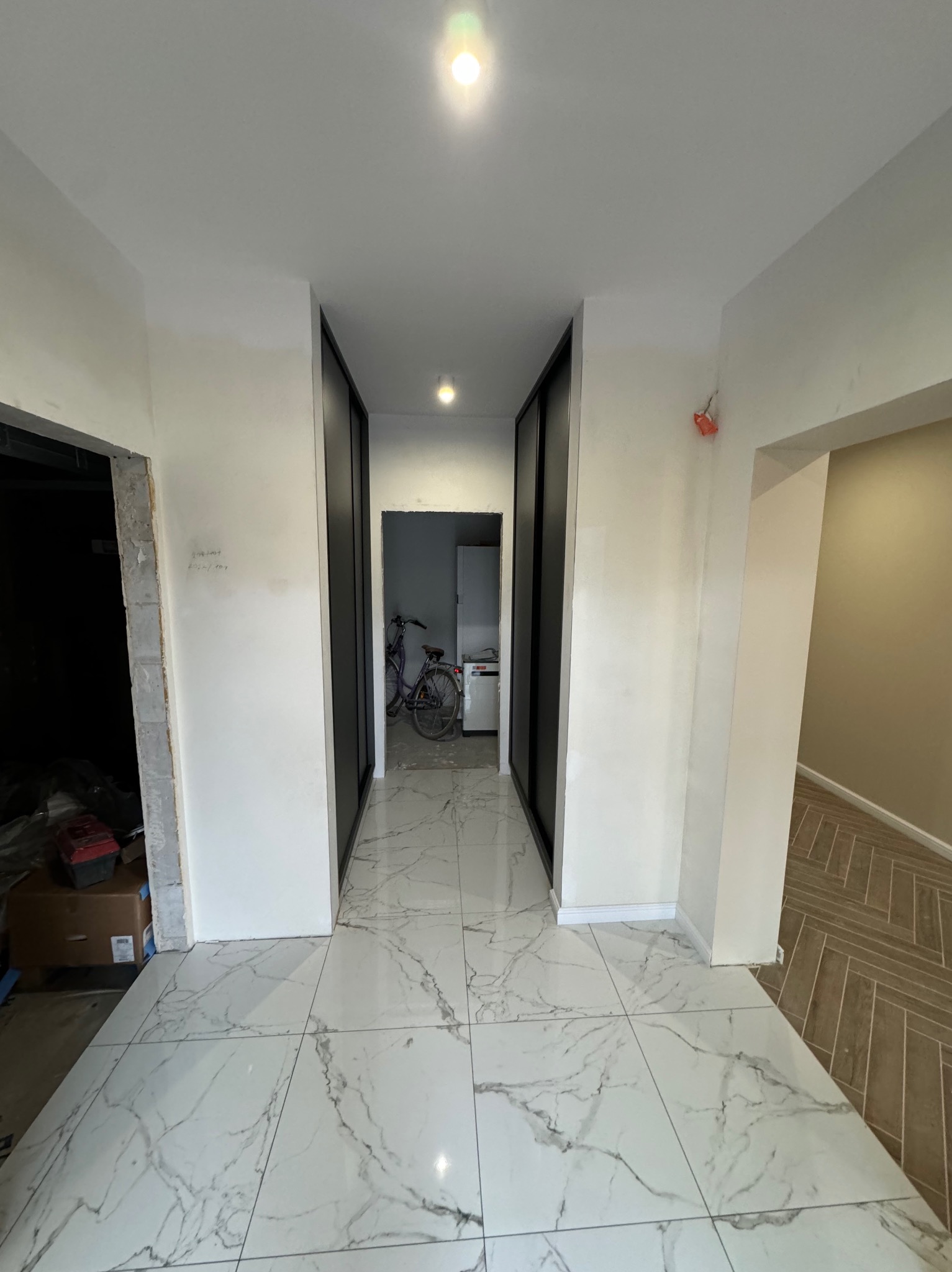 Korytarz w trakcie remontu z białymi ścianami, marmurową podłogą i widocznymi elementami instalacji elektrycznej. W głębi widoczny rower i szafa z przesuwnymi drzwiami.