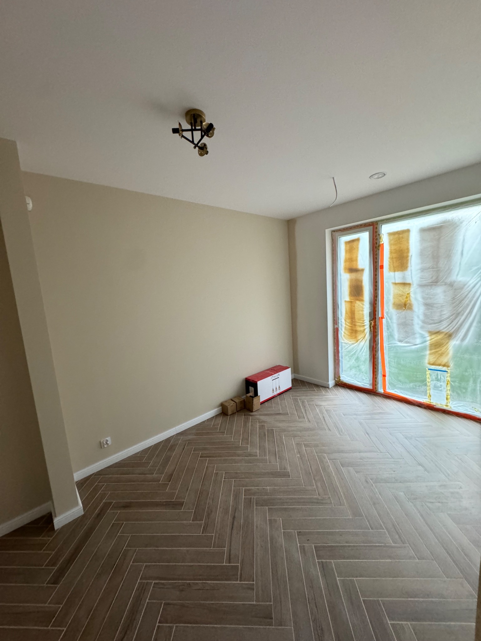 Pomalowane na beżowo wnętrze z parkietem ułożonym w jodełkę, okno zabezpieczone folią malarską, karton z lampą na podłodze.