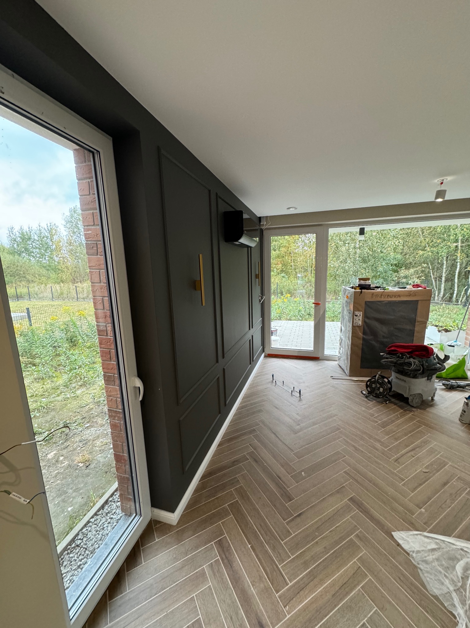 Wnętrze w trakcie prac wykończeniowych: ułożona podłoga z paneli w jodełkę, pomalowana na ciemno ściana z dekoracyjnymi listwami, widoczne narzędzia i materiały budowlane.