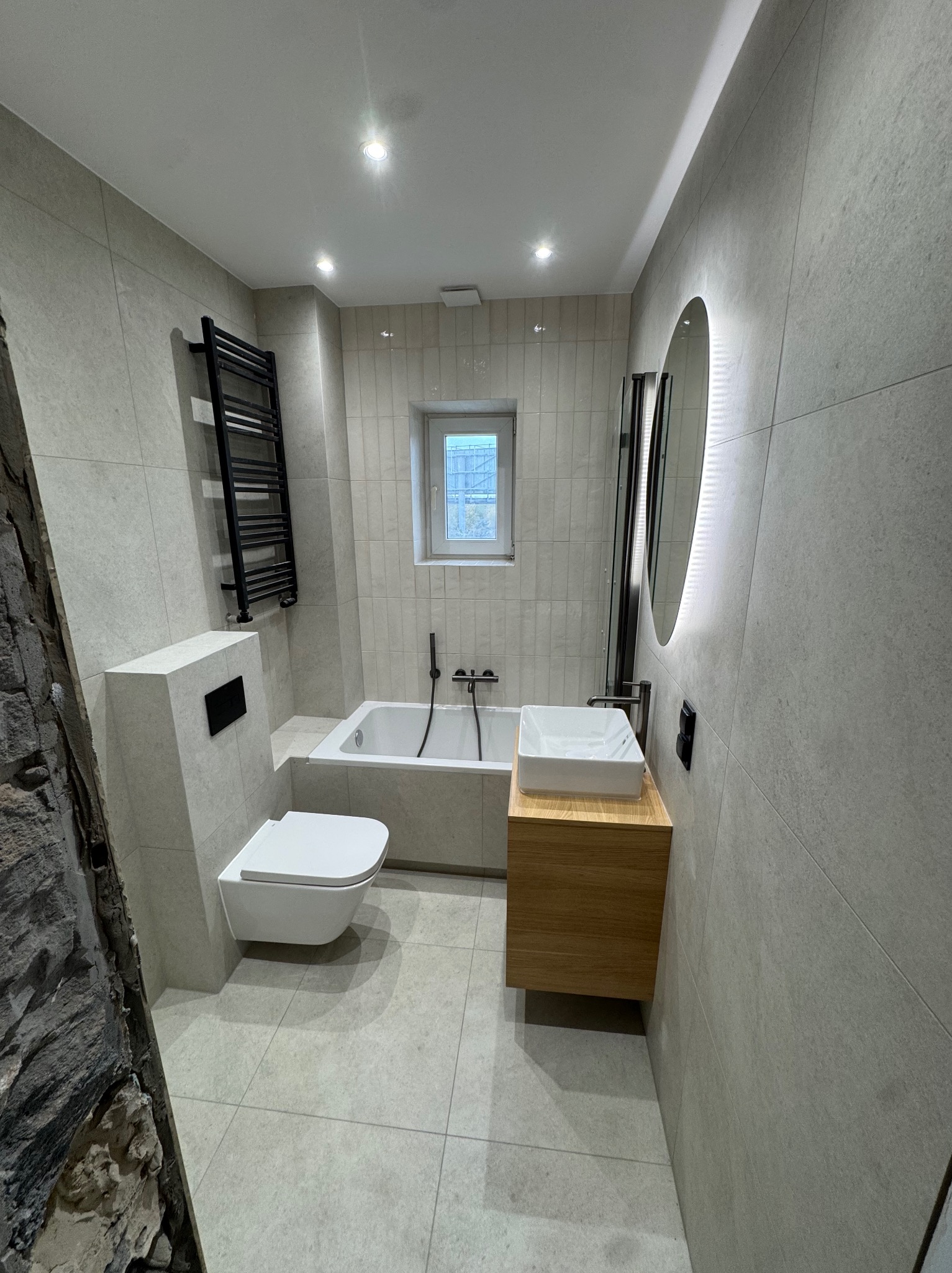 Nowoczesna łazienka z szarymi płytkami, podwieszaną toaletą, drewnianą szafką pod umywalką i okrągłym lustrem z podświetleniem LED. Widoczny fragment ściany z kamienia.