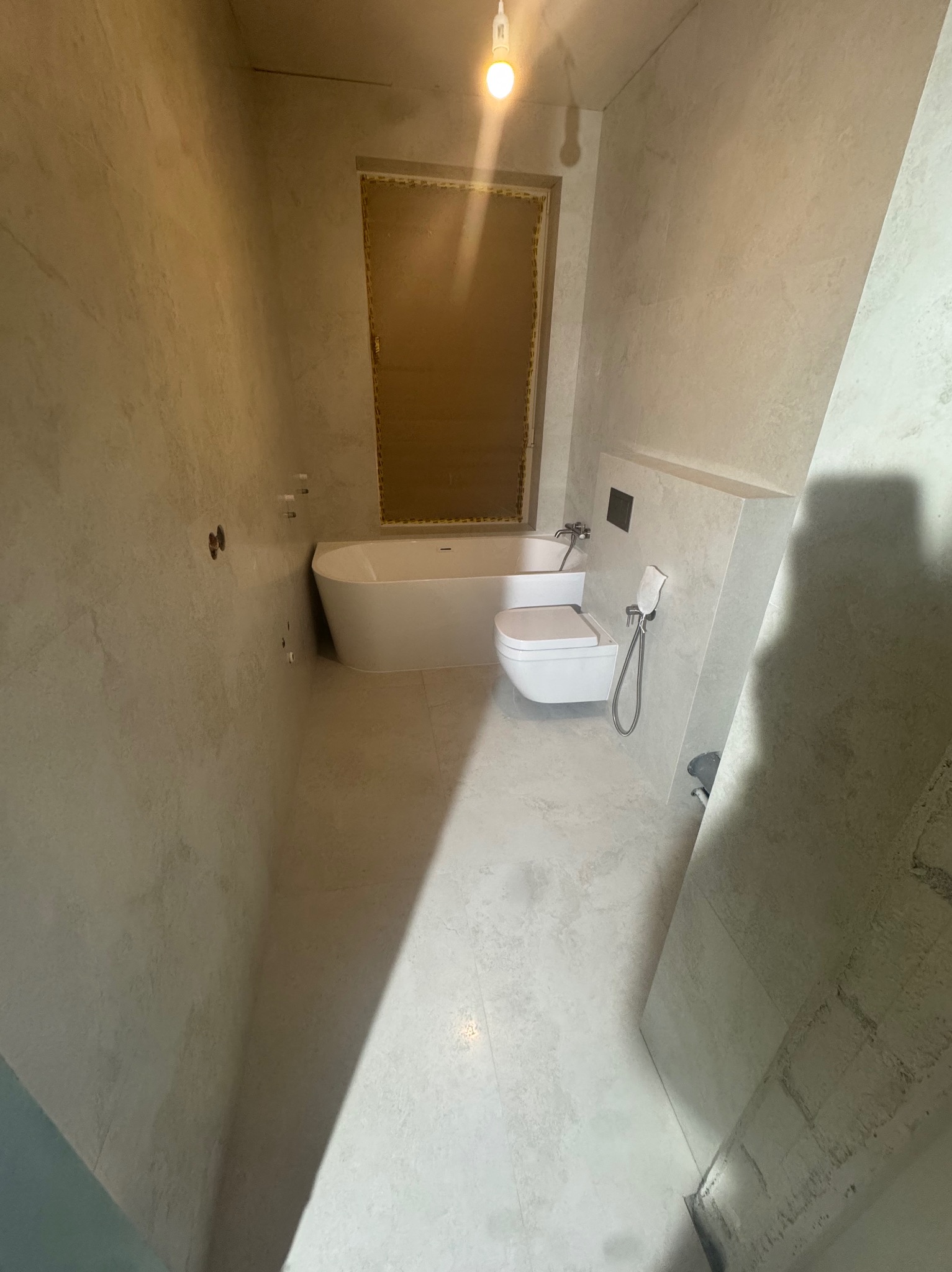 Jasna łazienka z wanną, toaletą i bidetką. Ściany wykończone w jasnym odcieniu z delikatną fakturą. Minimalistyczny design, surowy klimat, widoczne niedociągnięcia wykończeniowe.