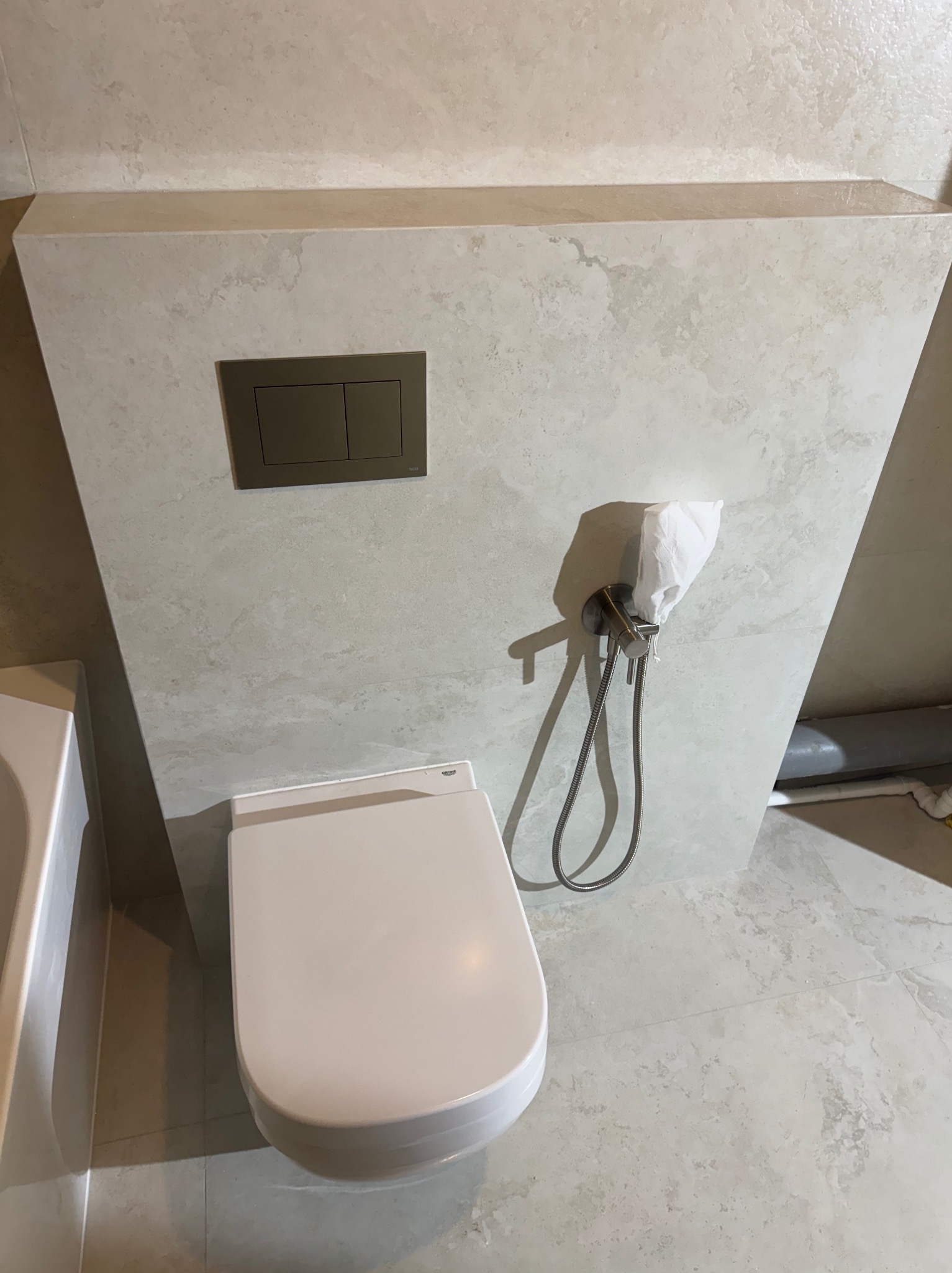 Nowoczesna toaleta podwieszana z szarym panelem sterującym i bidetką, wykończona płytkami imitującymi kamień. Minimalistyczny design łazienki.