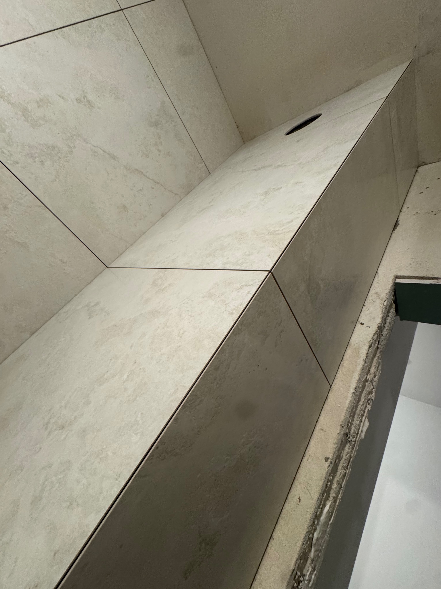 Ściana wyłożona beżowymi, matowymi płytkami ceramicznymi z ciemnymi fugami. Widoczny otwór w płytce. Krawędź ściany z widocznym klejem i fragmentem surowej ściany.