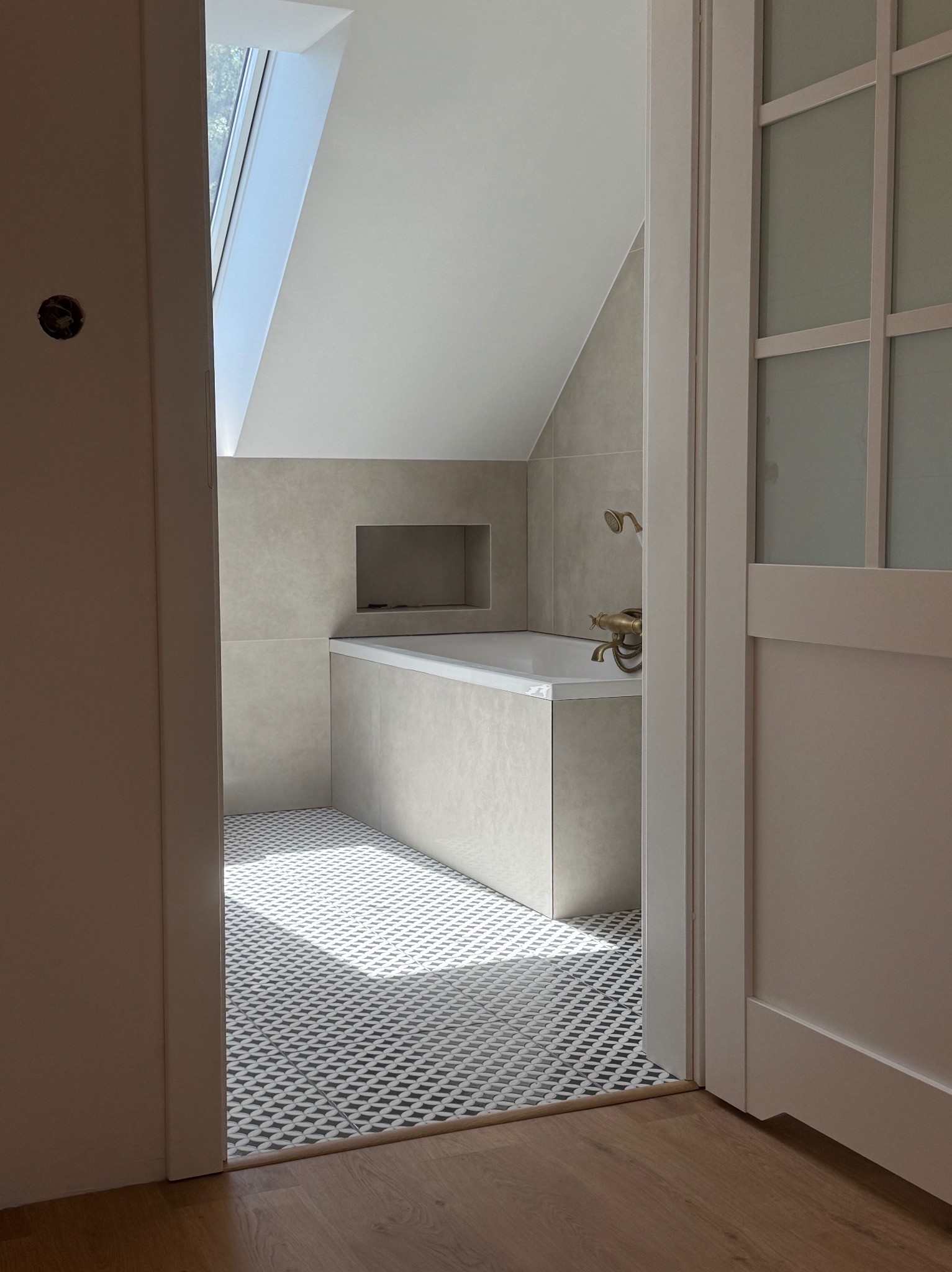 Wykończona łazienka z wanną pod skosem i oknem dachowym, widoczna przez otwarte drzwi. Na podłodze wzorzyste płytki, na ścianach jasne, duże kafle. Bateria wannowa w stylu retro.