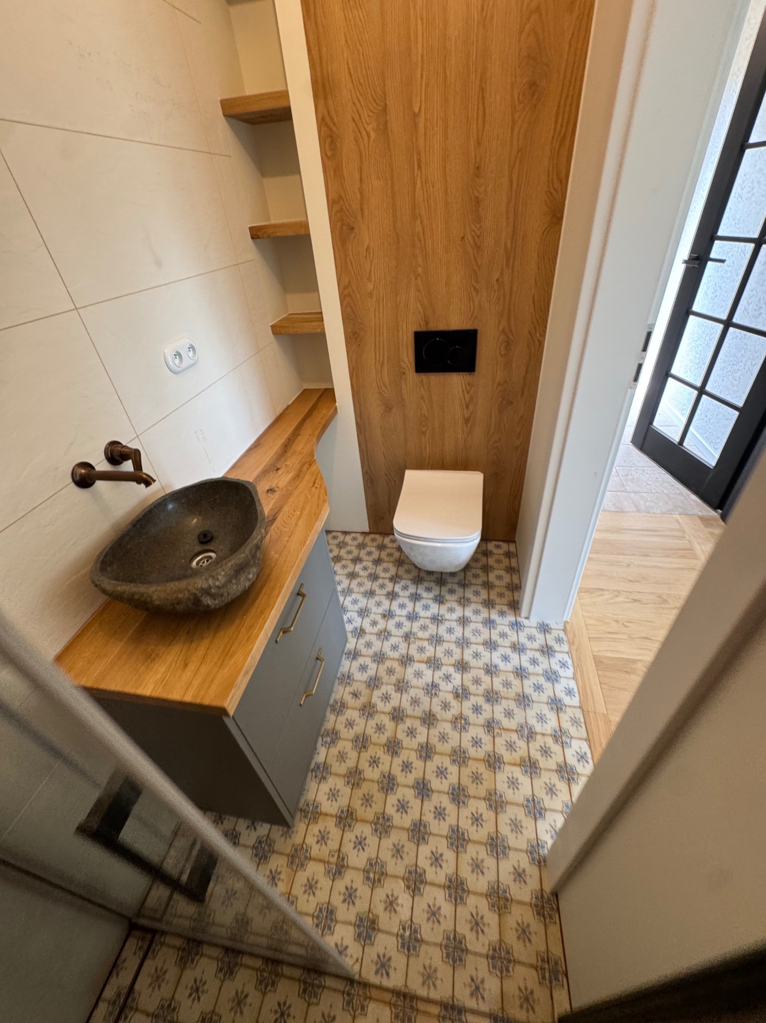 Nowoczesna łazienka z kamienną umywalką, drewnianym blatem i panelami ściennymi. Uwagę przykuwają wzorzyste płytki podłogowe i minimalistyczna toaleta.