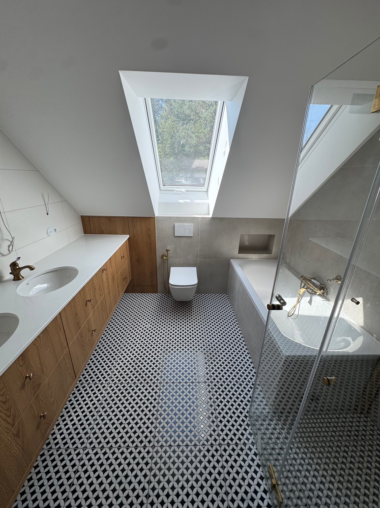 Jasna łazienka na poddaszu z oknem dachowym, wykończona w bieli, szarości i drewnie. Uwagę przykuwają wzorzyste, czarno-białe płytki podłogowe i złota armatura.