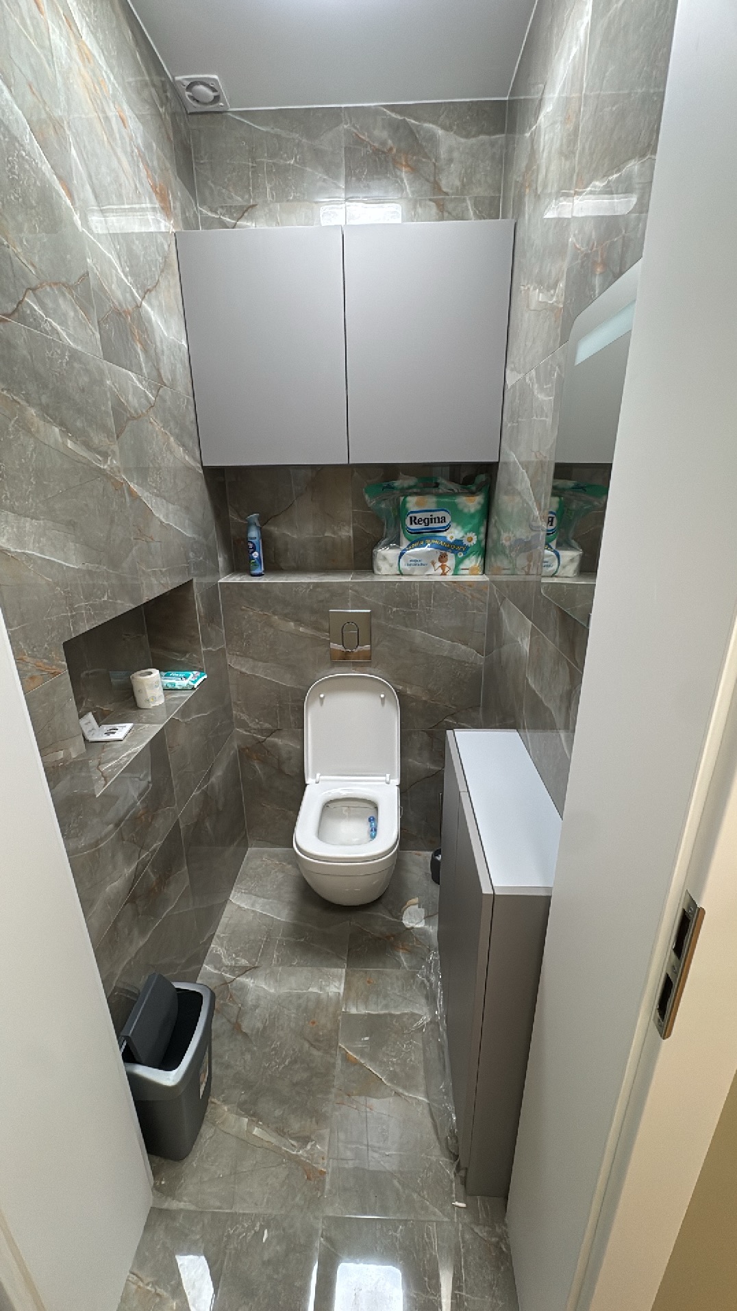 Nowoczesna toaleta z szarymi płytkami imitującymi marmur, podwieszana szafka nad WC i wnęka z papierem toaletowym. Widoczny kosz na śmieci i fragment szafki pod umywalkę.
